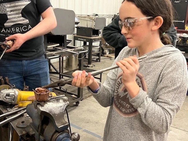 A young maker wearing safety glasses shaping a copper rose at a workshop vise.