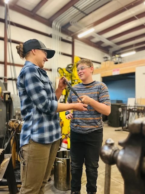 An instructor in a blue plaid shirt helping a smiling young maker with his metal project at Kilroy’s Workshop.